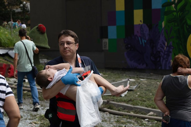 Socialni delavec nosi otroka, medtem ko reševalci, bolnišnično osebje in prostovoljci odstranjujejo ruševine in iščejo ljudi, ujete pod razvalinami po napadu na bolnišnico Okhmatdyt, največji otroški zdravstveni center v Ukrajini.
