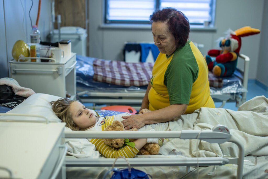 Deklica leži v postelji v bolnišnici, poleg nje sedi babica.