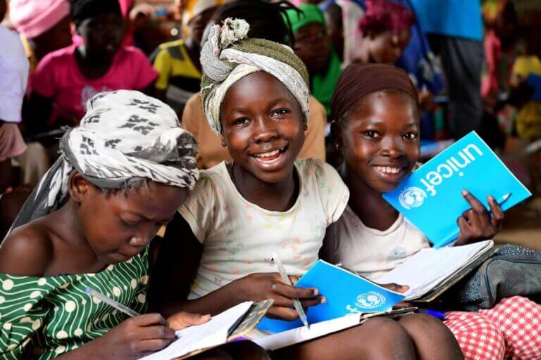 Tri nasmejane deklice, ki v rokah držijo UNICEF-ove pripomočke za šolo.