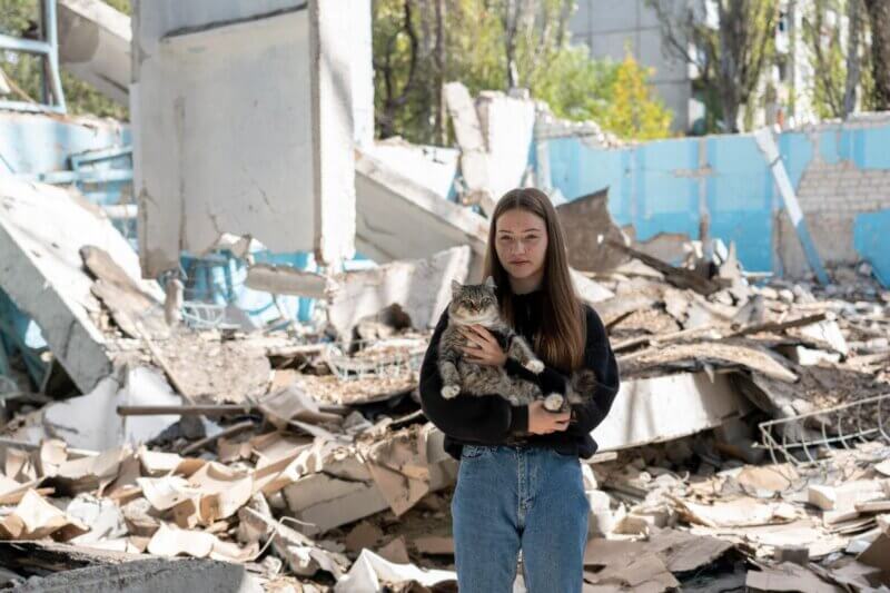 Deklica z mačko v naročju stoji med ruševinami šole.