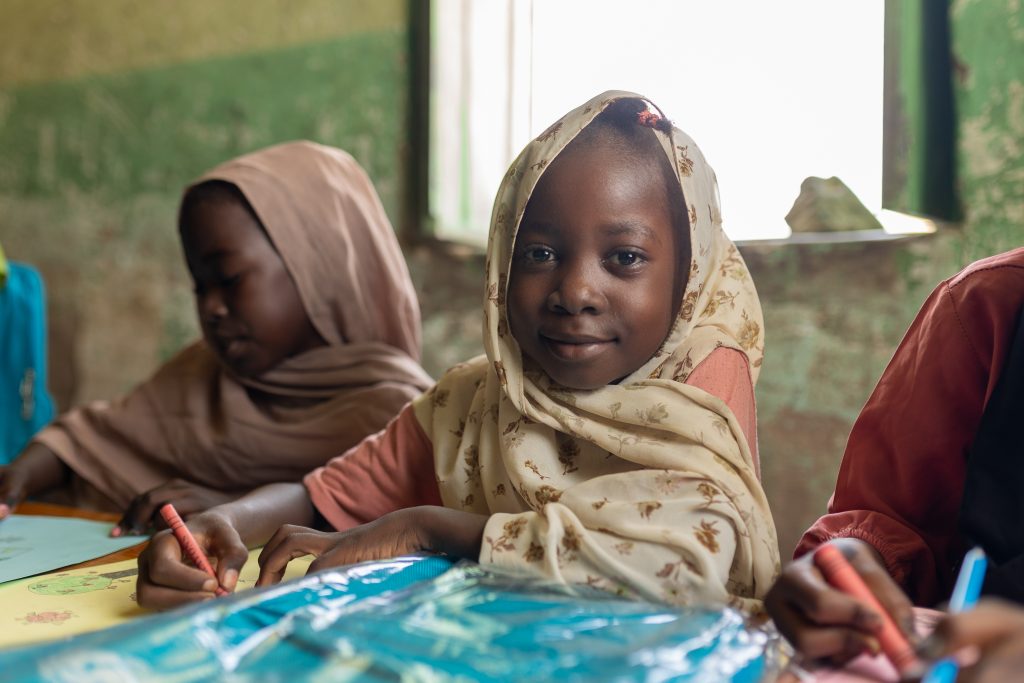 Deklica v šoli v Sudanu piše v UNICEF-ov zvezek in se nasmiha kameri.
