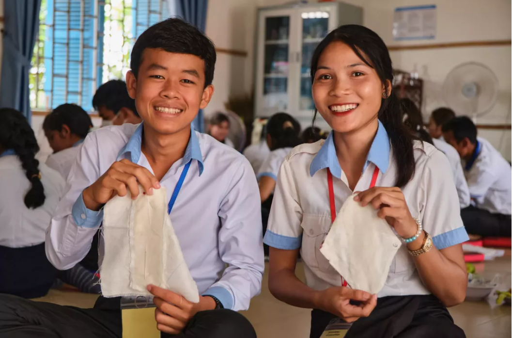 Učenci iz Kambodže držijo menstrualne higienske pripomočke in se smejijo kameri.