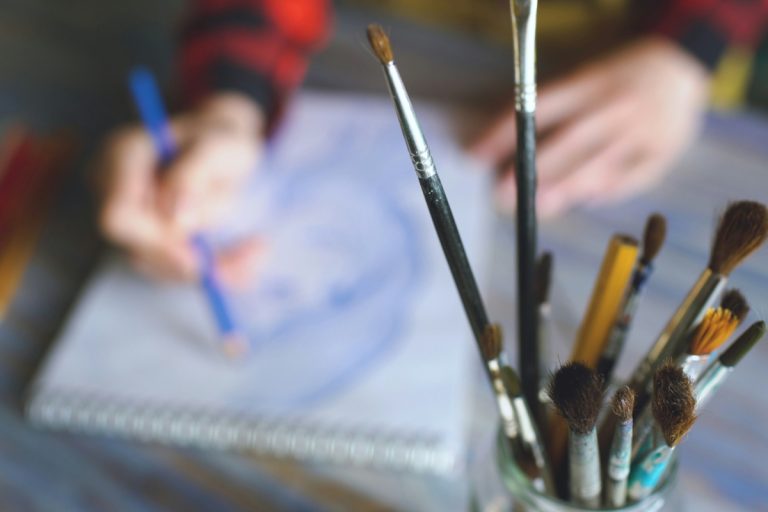 Otrok riše v skicirko z umetniškimi potrebščinami v bližini.