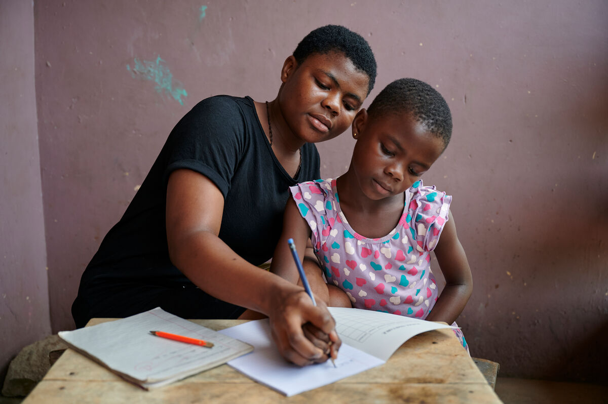 Sestrična Vera pomaga 7-letni Bennedikti pri domači nalogi pred hišo v občini Juaben v regiji Ashanti v Gani, Zahodna Afrika.
