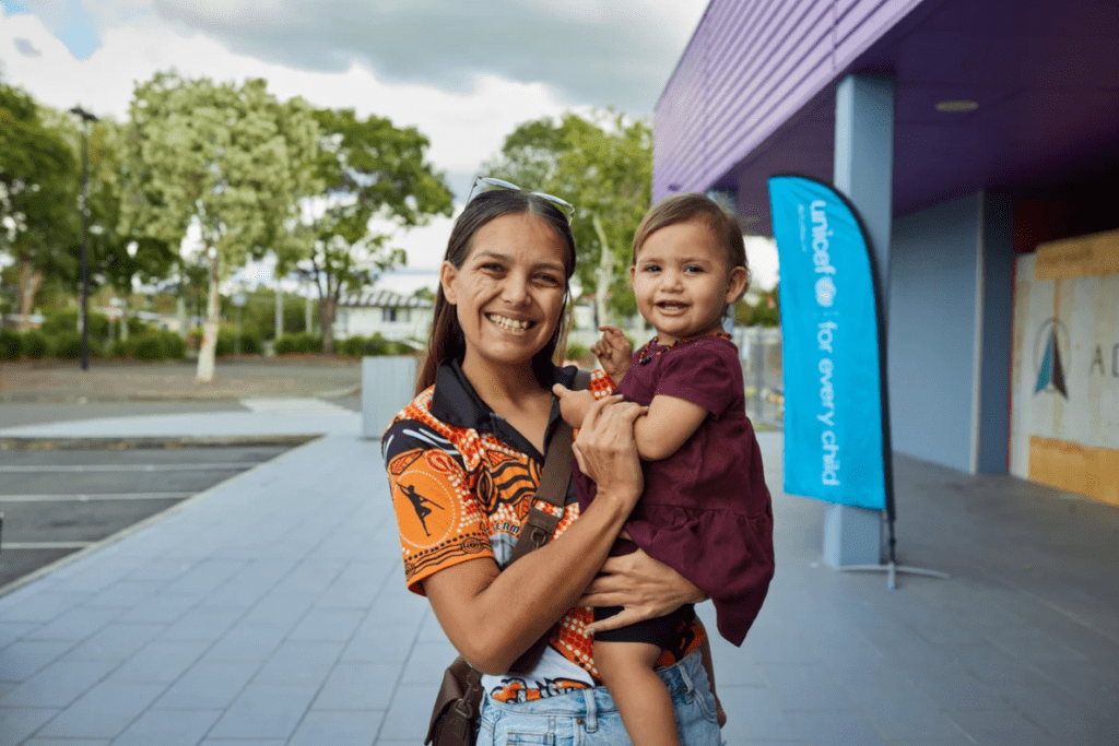 Mati drži hčerko v roki pred UNICEF-ovim centrom in se smejita kameri.