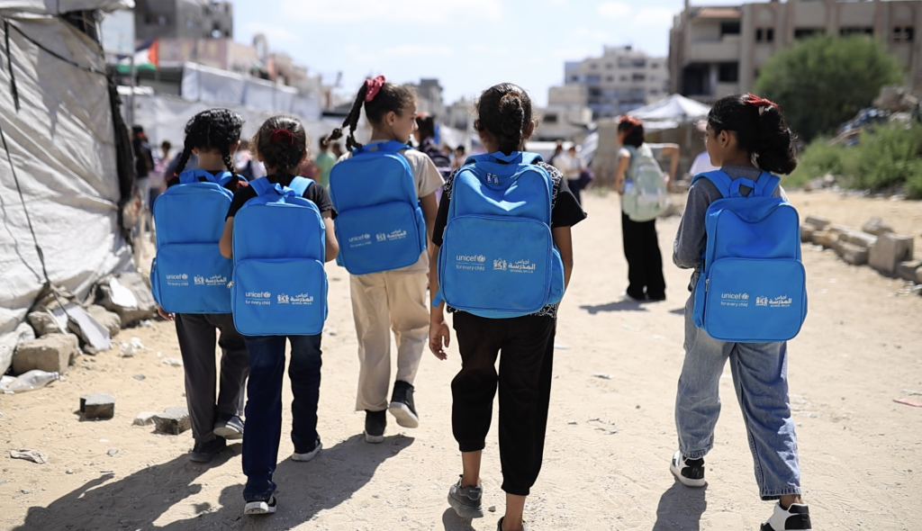 Učenci z UNICEF-ovimi torbami hodijo po ulici v Gazi, v ozadju so vidne ruševine in posledice uničenja.