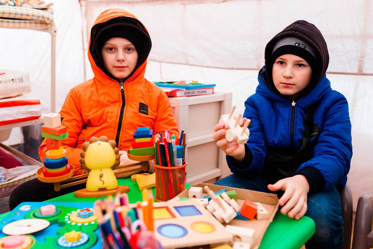 Dva otroka v Modri točki, pred njima je miza z igračami in pisali.