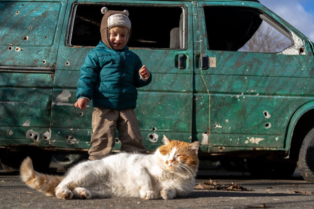 3-letni Jehor iz Ukrajine stoji pred preluknjanim kombinjem in se smeji svojemu mačku Frenku, ki sta ga z mamo rešila iz ulice.