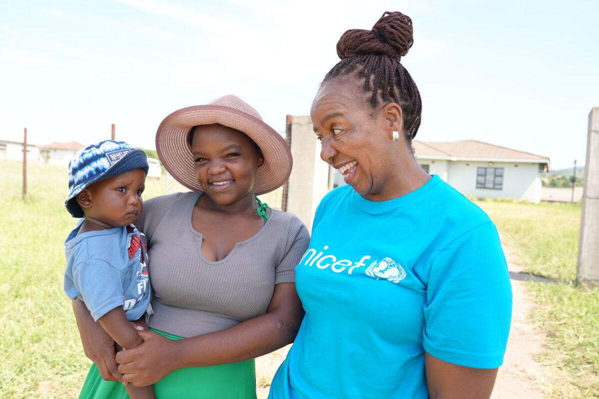 Mati drži svojega otroka v roki in skupaj z UNICEF-ovo sodelavko se smejijo kameri.