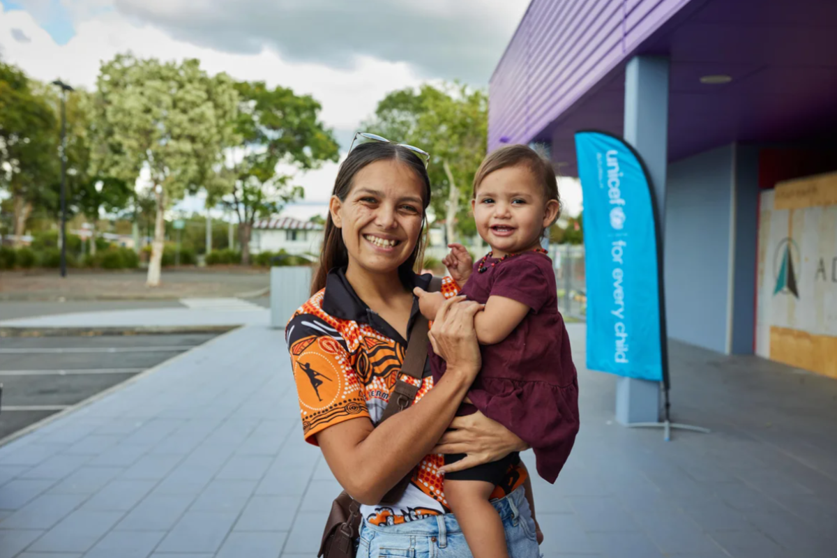 Mati drži hčerko v roki pred UNICEF-ovim centrom in se smejita kameri.